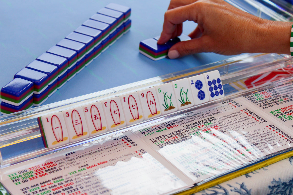A close-up of a hand playing with the coastal chic La Jolla Tiles Mahjong set, placing a four layer acrylic tile onto blue coastal chic mat with score card visible.